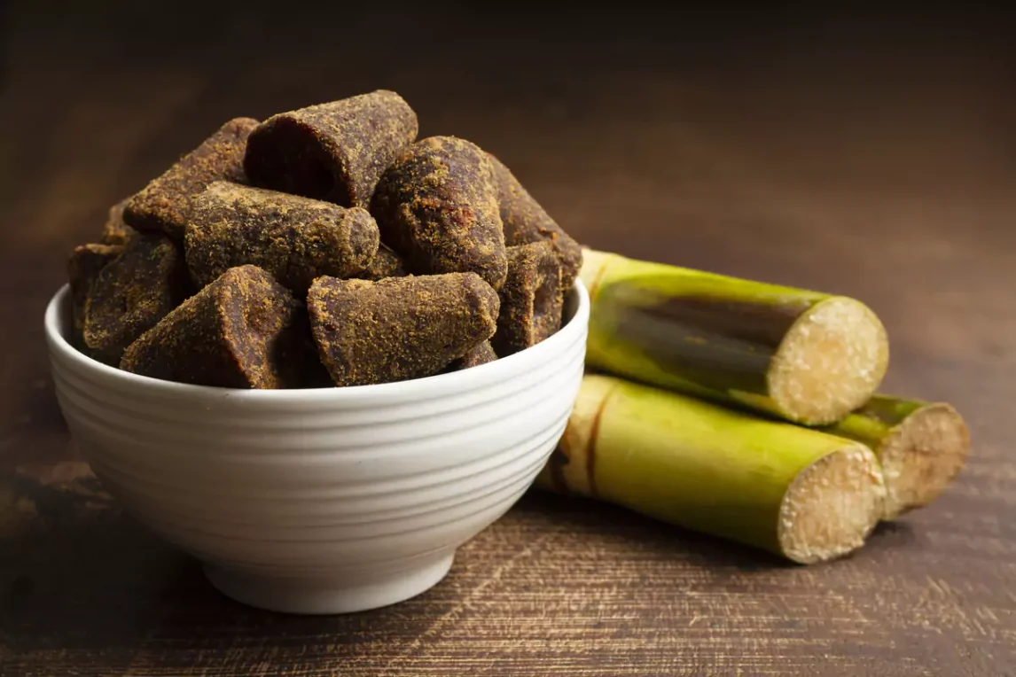 Traditional Mexican piloncillo cones stacked in a bowl next to some sugar cane, highlighting their deep brown color and unrefined texture.