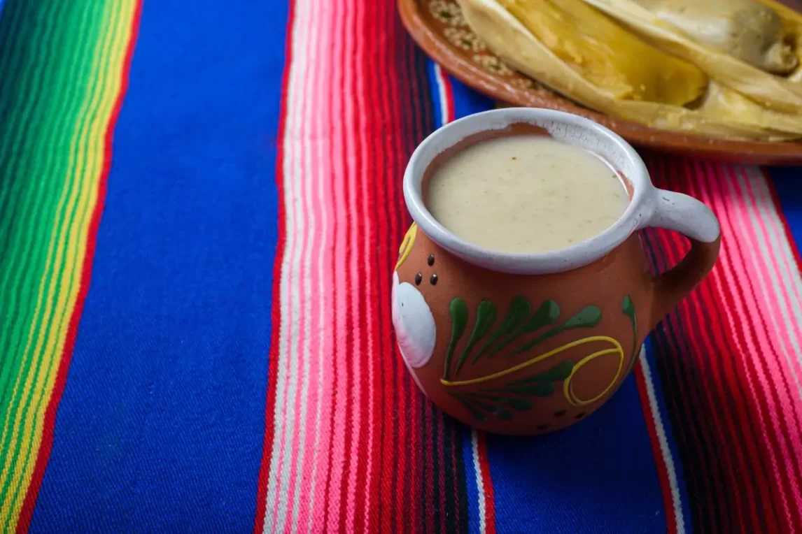 Traditional clay mug filled with creamy blue corn atole on a vibrant striped Mexican textile, served alongside tamales for a warm, comforting Southwest meal.