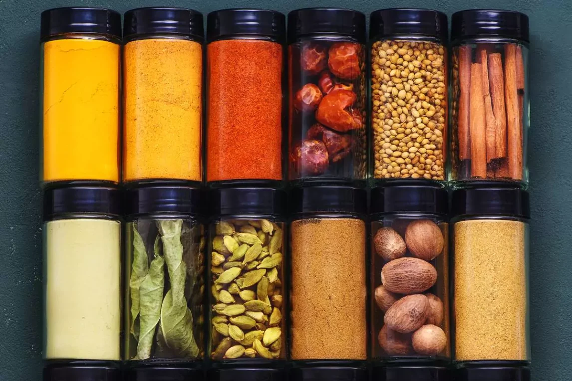 A colorful lineup of neatly organized spice jars including cinnamon sticks, dried chiles, and peppercorns used to illustrate spice shelf life and storage tips.