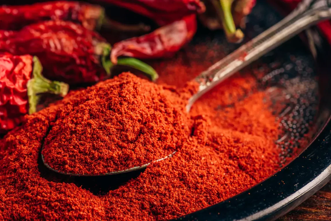 Close-up of vibrant red chile powder in a spoon with dried red chiles in the background, showcasing rich texture and bold Southwest spice.