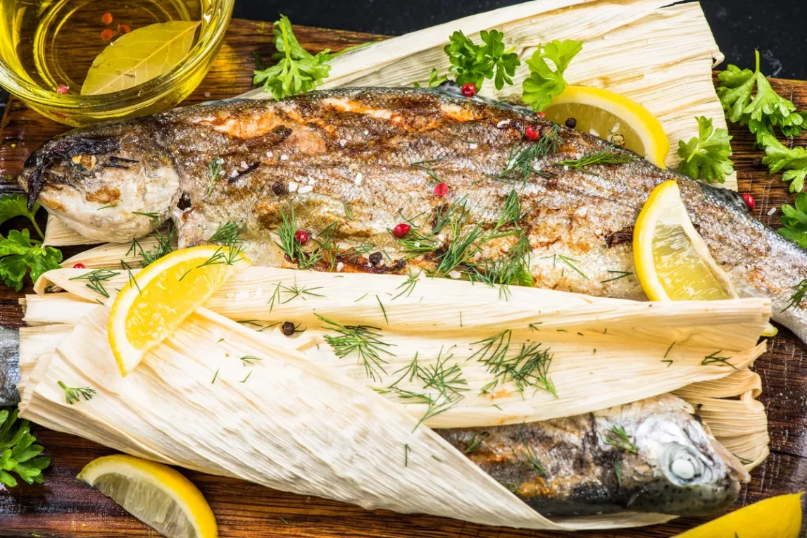 Grilled whole fish wrapped in corn husks with lemon, herbs, and spices, showing a traditional method of cooking with corn husks.