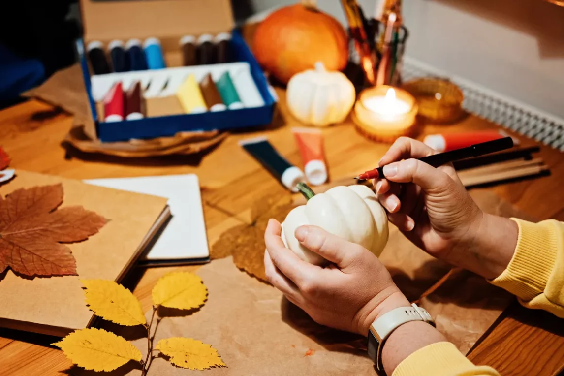 Hands painting a small white pumpkin on a fall craft table with leaves, paints, and candles, showcasing typical Thanksgiving crafts.