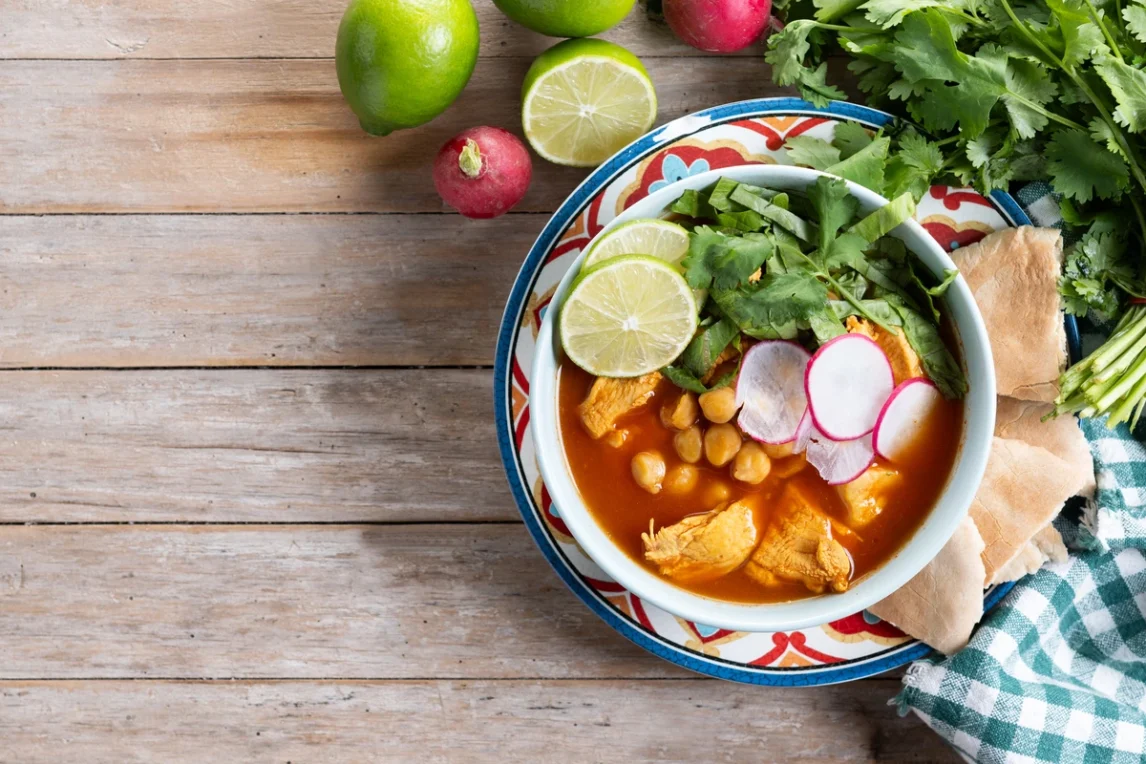 A bowl of traditional New Mexican red chile posole with chicken, hominy, lime, cilantro, and radish slices on a rustic wooden table.