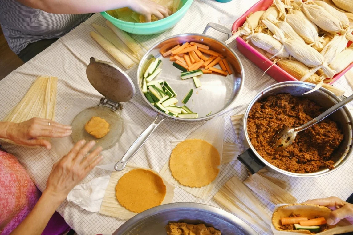 Hands preparing tamales during a tamalada, with corn husks, masa, vegetables, and seasoned meat filling spread across the table for a holiday gathering.